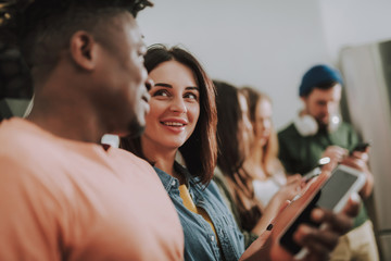 Obraz premium Portrait of beautiful girl gazing at friend and smiling while he holding smartphone. Bearded gentleman and two women on blurred background