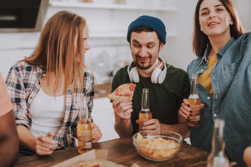 Portrait of bearded man with funny facial expression holding slice of pizza and bottle of beer while sitting at the table with two girls