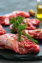 Raw pork cheeks meat on a dark gray surface with rosemary and spices.