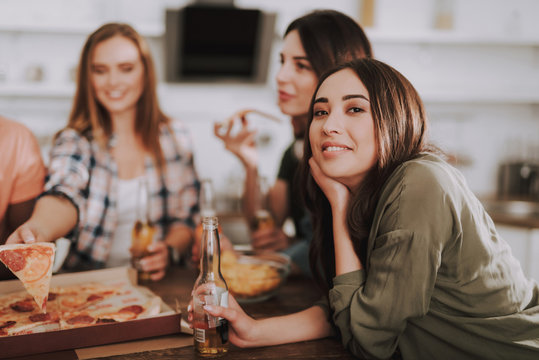 Portrait of charming girl with bottle of beer looking at camera and smiling while sitting at the table. Two women with pizza on blurred background