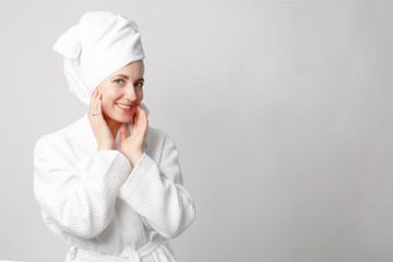 Beautiful girl wearing white bathrobe with towel on the head, looking at camera and smiling, a model with light nude make-up, white studio background, beauty photo, copy space.