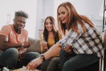 Portrait of two charming girls and handsome afro american guy sitting on couch and holding bottles of beer