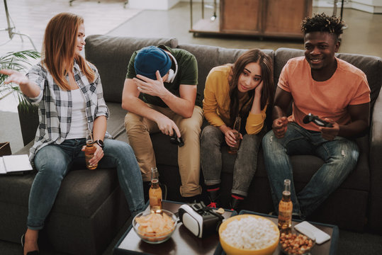 Game over. Portrait of young man in hat covering face with hand while afro american guy smiling. Girls sitting on couch and feeling disappointed