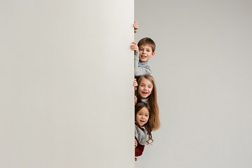 Banner with a surprised children peeking at the edge with copyspace. The portrait of cute little kids boy and girls looking at camera against white studio wall. Kids fashion and happy emotions concept
