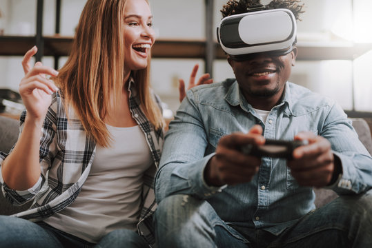 Portrait of joyful afro american guy wearing virtual reality glasses and holding joystick while young lady looking at him and laughing. They sitting on couch at home