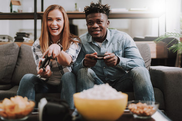 Portrait of beautiful girl and afro american guy sitting on couch and using joysticks. They are smiling