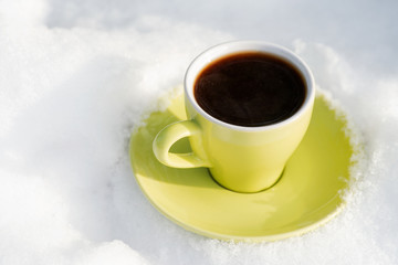 Winter holiday decoration concept: green cup of coffee with saucer on frozen snow covered wood log in forest preserve park.