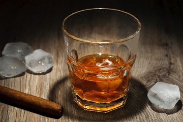 A glass of alcoholic beverage with ice cubes and a cigar on a wooden table. Whiskey in glass