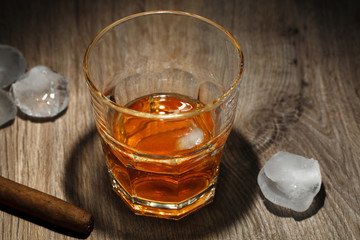 A glass of alcoholic beverage with ice cubes and a cigar on a wooden table.