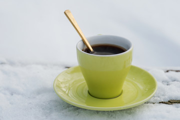 Winter holiday decoration concept: green cup of coffee with saucer on frozen snow covered wood log in forest preserve park.