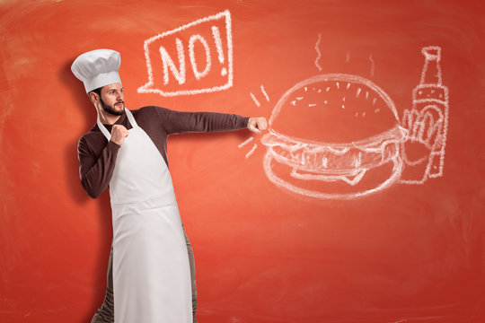 A Young Handsome Chef Saying 'No' And Hitting With Clenched Fists A Chalk Drawing Of A Hamburger, French Fries And A Ketchup Bottle.