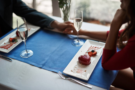 Concept of lovely and romantic date. Top angle of beloved couple holding hands while sitting in restaurant with sweet dessert and champagne