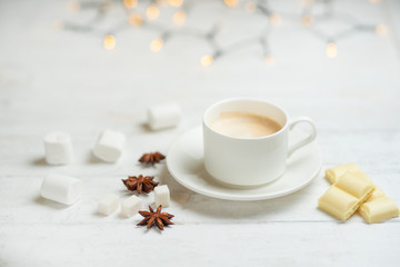 Obraz na płótnie Canvas Coffee with milk, latte with cinnamon sticks and anise stars with white chocolate and marshmallow, on a light white background, with lights from a garland.