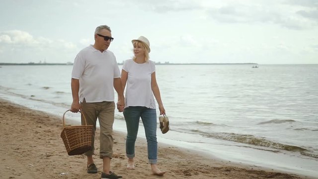 Senior lovers walking and takling on shoreline after beach picnic. Mature woman in straw hat and with bare feet holding hand of happy retired man.
