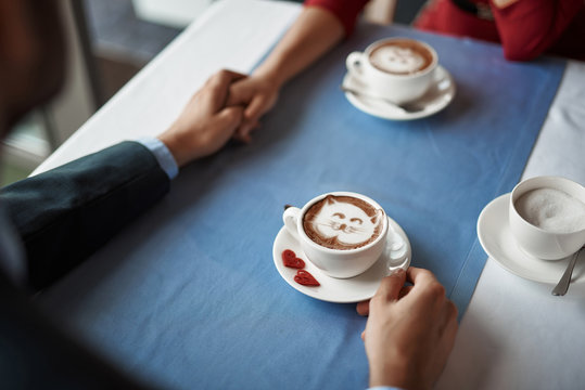 Concept of lovely date. Top view sweet cup of coffee with cat picture and hearts decore staying on table where sitting beloved couple and holding hands