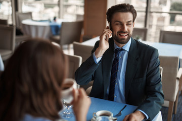 Business lunch in friendly atmosphere. Close up selective focus on smiling businessman talking by phone while sitting in restaurant with woman that drinking coffee