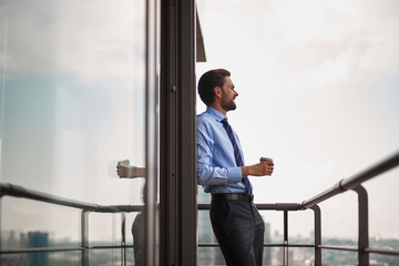Corporate time-out culture. Low angle side on portrait of businessman standing on office balcony with cup of coffee and looking at city view