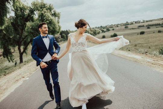 Wedding Couple Running On The Road.Happy Young Bride And Elegant Groom Running Away Holding Hands.Stylish Couple Of Newlyweds On Their Wedding Day. 