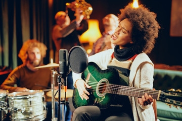 Mixed race woman singing and playing guitar while sitting on chair with legs crossed. In background...