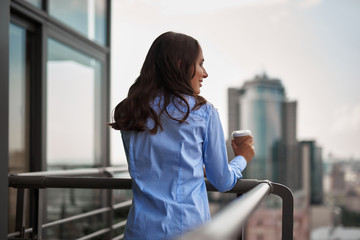 Corporate time-out. Waist up back side portrait of young smiling office woman standing on balcony with cup of coffee
