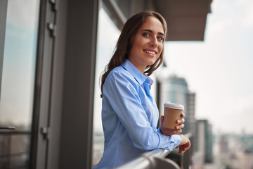 Corporate time-out. Waist up portrait of young smiling office woman standing on balcony with cup of coffee. Copy space on right