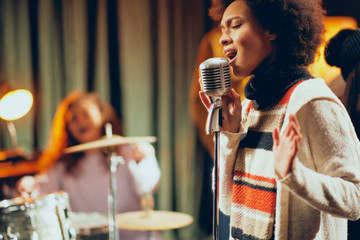 Close up of mixed race woman singing. In background band playing instruments. Home studio interior.