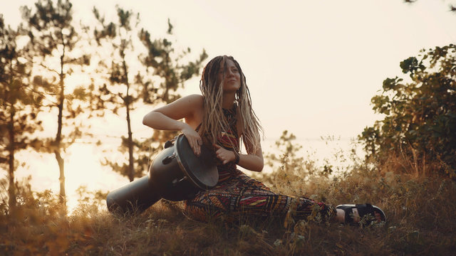 Beautiful Young Hippie Woman With Dreadlocks Playing On Djembe. Funky Woman Drumming In Nature On An Ethnic Drum At Sunset Or Sunrise