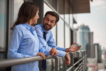 Concept of friendly communication between coworkers. Portrait of young smiling man and woman in office clothes having conversation on balcony while have work break - obrazy, fototapety, plakaty