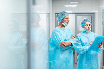 Calm serious doctor standing with his arms crossed and thoughtfully looking at the notes in the hands of his younger colleague. Copy space on left side