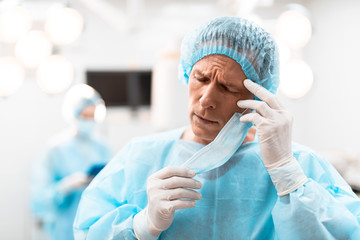 Exhausted doctor having headache and closing his eyes while putting off the mask and touching his forehead