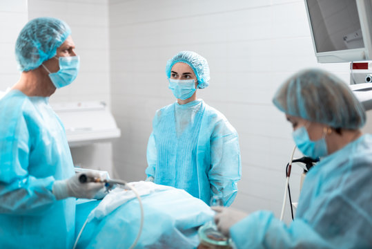 Waist up portrait of female medical worker in sterile gown staring at doctor. He holding laparoscopic instrument and checking patient condition on monitor. Focus on girl