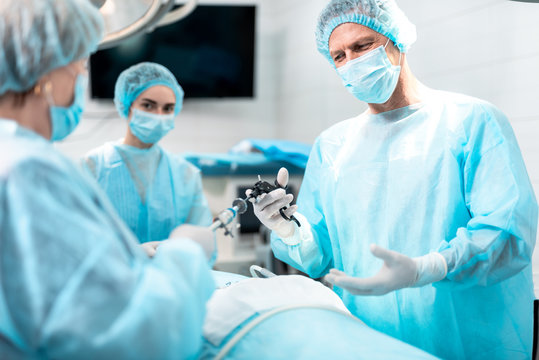 Medical team. Waist up portrait of smiling doctor in protective mask holding laparoscopic grasper with trocar while standing in operating room with his assistants