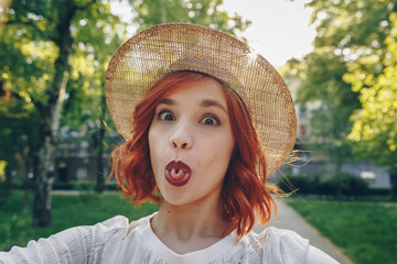 Beautiful and stylish red haired girl taking a selfie with her smartphone outdoors, authentic...