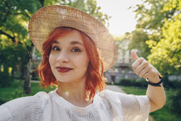 Beautiful and stylish red haired girl taking a selfie with her smartphone outdoors, authentic...