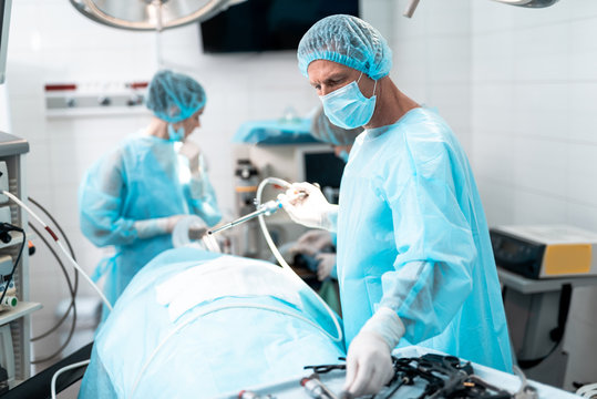 Waist up portrait of middle aged doctor in protective mask performing surgery in modern operating room. Female assistant on blurred background