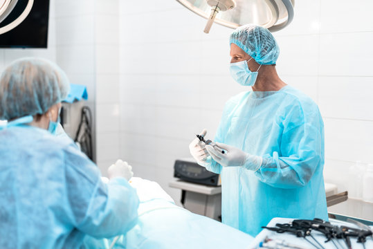 Portrait of doctor in sterile gloves looking at surgical table with patient before surgery. He is wearing blue cap and gown