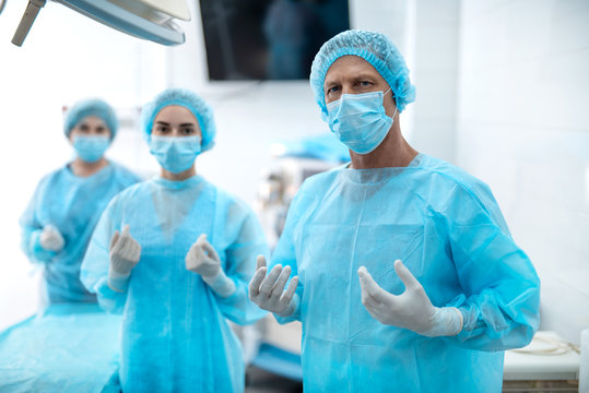 Best team. Waist up portrait of surgeon in protective mask and sterile gloves looking at camera with serious expression while his assistants standing behind him on blurred background