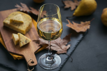 A glass of white wine served with cheese in a cutting board on dark background