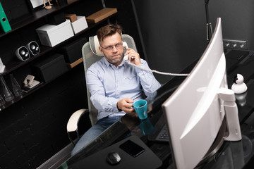 man leaned back in his chair, drinking coffee and talking on landline