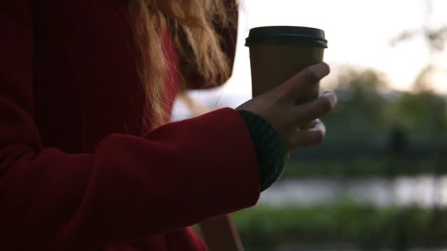 Young woman in red coat is drinking coffee on the street while walking on cold autumn day. Close-up of hands with brown take away cup of hot coffee. Red head woman walking by city rainy park
