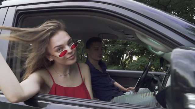 Young couple travels by car Lady in red shirt and sunglasses shows her tounge Man is driving vehicle