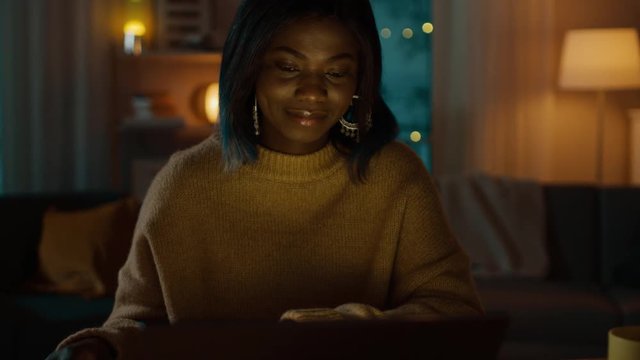 Portrait of Beautiful Smiling Black Girl Working on a Laptop while Sitting at Her Desk at Home. In the Evening Creative Woman Works on a Computer In Her Cozy Living Room. 