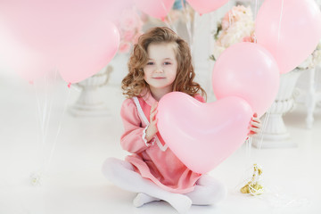 Small girl with blond curly hair poses in a white studio surrounded by pink balloons. A beautiful preschool girl in a white studio with heart-shaped pink balloons. Valentine's Day, love, birthday.