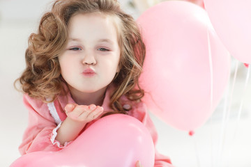 Small girl with blond curly hair poses in a white studio surrounded by pink balloons. A beautiful preschool girl in a white studio with heart-shaped pink balloons. Valentine's Day, love, birthday.