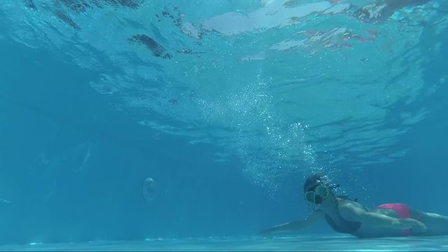 girl in a mask under the water in the swimming pool looks into the camera, bubbles, sun rays, outdoors