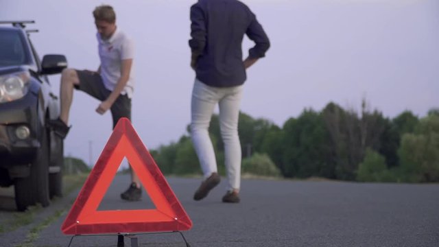Broken black car is standing at the road Man in white jeanse puts warning triangle on the asphalt Car trouble Focus on the foreground