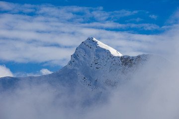 Distant landscape of snowy mountain peak with clouds reaching you on a cold afternoon of blue sky.
