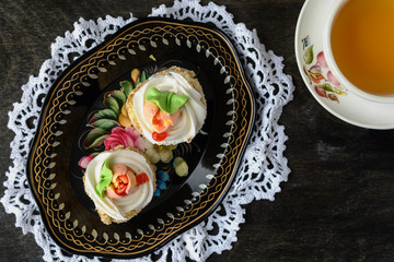 Two biscuit cakes with butter cream, decorated with flowers from the cream are on a beautiful vintage metal tray and white lace doily are next to a cup of tea on a dark background, top view