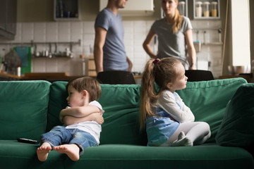 Upset offended toddler brother and sister sitting separately on couch, sofa with arms crossed,...