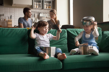 Happy toddler girl and boy playing with kitchenware on sofa at home, little sister and brother laughing using pots as drums and hats, holding kitchen utensils, making noises, spending time together